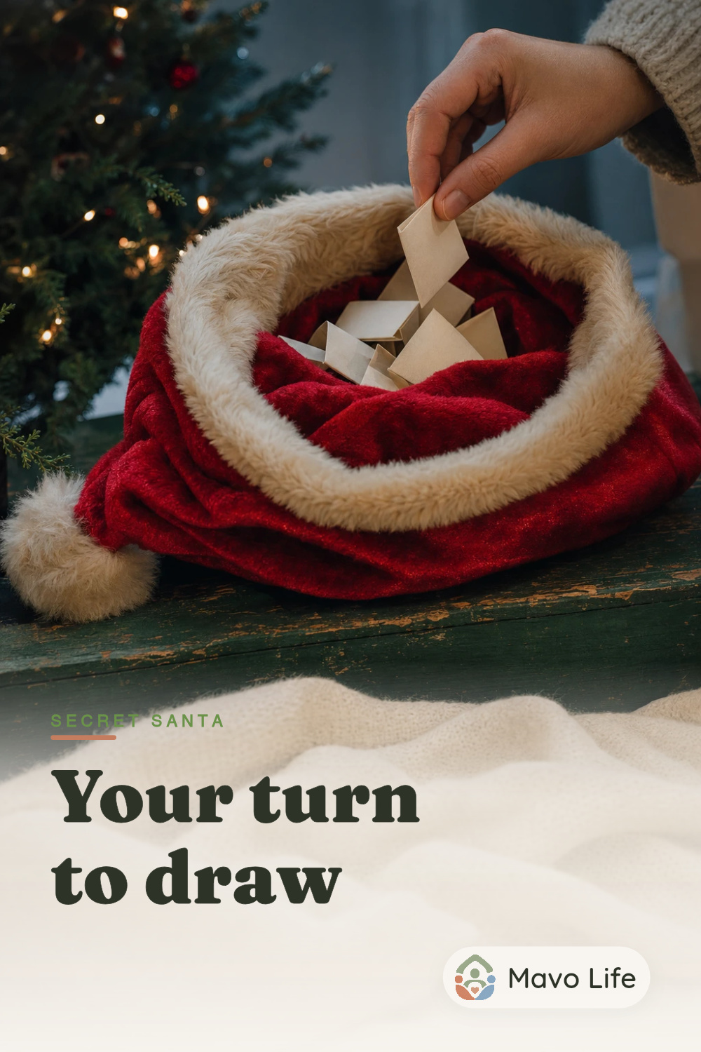 An illustrative traditional Secret Santa draw: a hand lifts one folded name slip from a red velvet Santa hat beside a Christmas tree. Text reads Secret Santa, Your turn to draw.